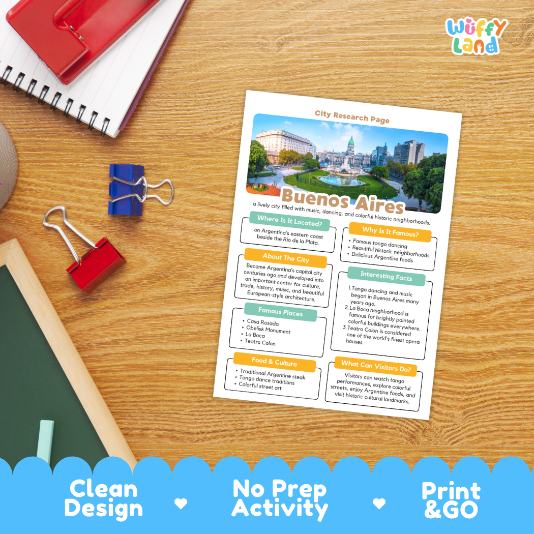 A printed city research page about Buenos Aires lies on a wooden desk, surrounded by school supplies including a red notebook, binder clips, a chalkboard, and a blue pen. The page has a colorful city photo at the top and text sections about the location, food, and culture.