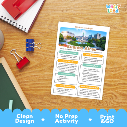 A printed city research page about Buenos Aires lies on a wooden desk, surrounded by school supplies including a red notebook, binder clips, a chalkboard, and a blue pen. The page has a colorful city photo at the top and text sections about the location, food, and culture.