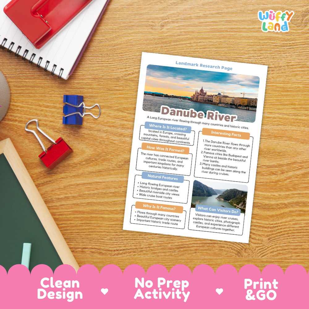 A printed Danube River landmark research page laid on a wooden desk, surrounded by school supplies including a red notebook, blue binder clips, a green chalkboard, and a red folder. The page has a clean design with text sections and small photos.
