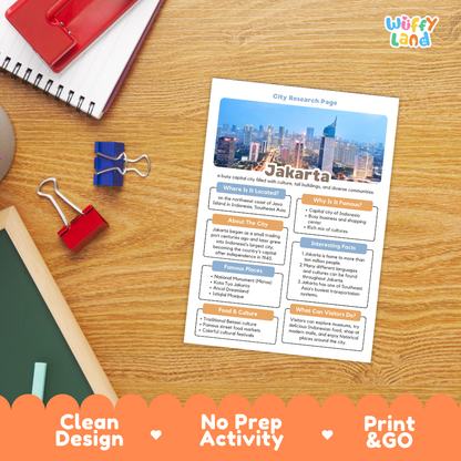 A printed city research page about Jakarta is shown on a wooden desk with school supplies around it, including a spiral notebook, binder clips, and a chalkboard. The page has a colorful city skyline photo at the top and text sections about Jakarta.