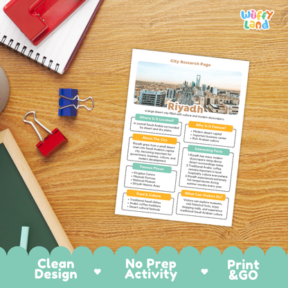 A printed city research page about Riyadh is displayed on a wooden desk with notebooks, binder clips, and a chalkboard nearby. The sheet has a colorful layout with headings, text blocks, and a city skyline photo.