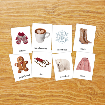Seven small printed cards with winter-themed illustrations and labels are arranged on a wood surface. The cards show mittens, a mug of hot chocolate with a heart-shaped drink, a snowflake, boots, a gingerbread figure, a sled, a polar bear, and a pink jacket.